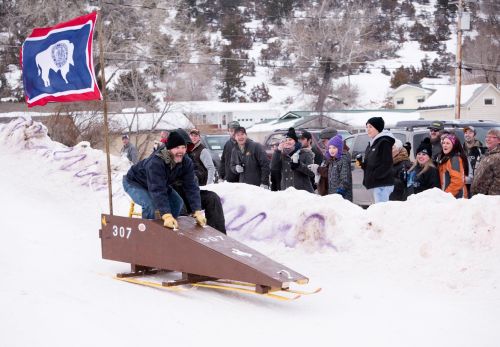 A person sleds down a snowy hill on a wooden sled, while a cheering crowd watches nearby. A flag waves in the background.