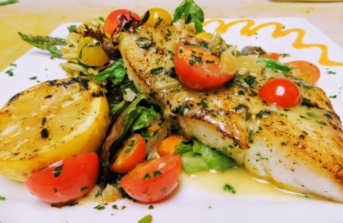 Grilled fish fillet topped with herbs, cherry tomatoes, and lemon, served on a bed of mixed greens and vegetables.