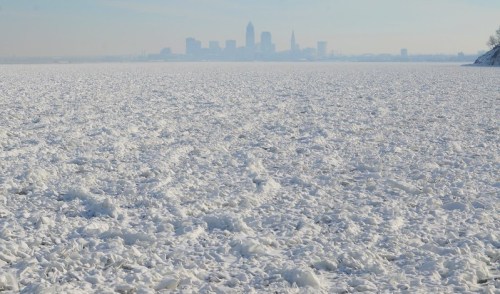 A frozen lake with ice formations, with a city skyline visible in the background under a clear sky.