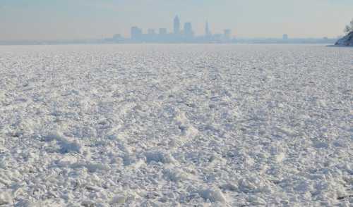 A frozen lake with ice formations, with a city skyline visible in the background under a clear sky.