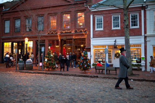 A festive street scene with decorated trees, holiday lights, and people shopping in a charming historic area.