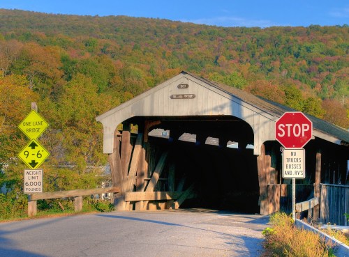 A covered bridge with a stop sign and weight limit signs, surrounded by colorful autumn foliage and hills.