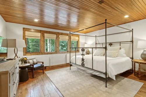 Cozy bedroom with a four-poster bed, wooden ceiling, large windows, and warm lighting.