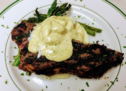 A plate featuring grilled steak topped with creamy sauce, served with asparagus and mashed potatoes, garnished with herbs.