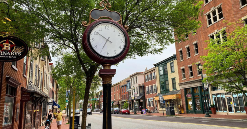 A vintage clock stands on a street lined with trees and historic buildings, with people walking in the background.