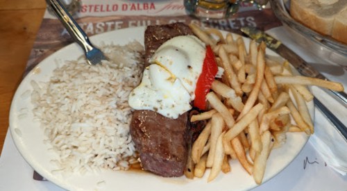 A plate of steak topped with a fried egg, served with rice and French fries, on a wooden table.