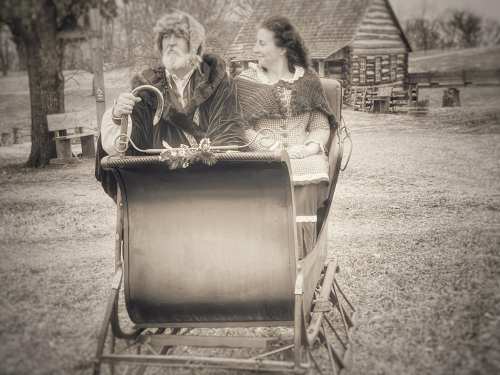 A vintage black-and-white photo of a man and woman in a horse-drawn sleigh, set in a rustic outdoor scene.