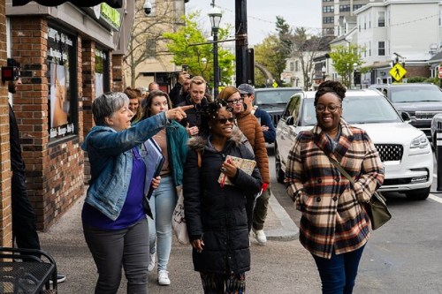 A diverse group of people walking together on a city street, smiling and chatting, with shops in the background.