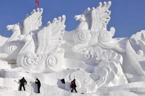 Snow sculptures of mythical creatures with wings, set against a clear blue sky, as people work nearby.