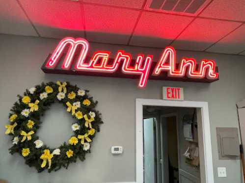 Neon sign reading "Mary Ann" above a wreath with yellow flowers and ribbons, next to an exit sign.