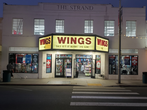 A brightly lit store named "WINGS" with a sale sign, featuring various items in the window, at dusk.