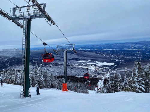 A snowy mountain landscape with ski lifts and gondolas, overlooking a valley and distant hills under a cloudy sky.