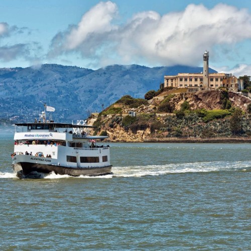 A ferry sails near Alcatraz Island, with mountains and clouds in the background.