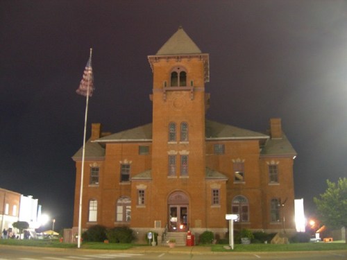 Historic brick building at night, featuring a tall clock tower and an American flag in front.