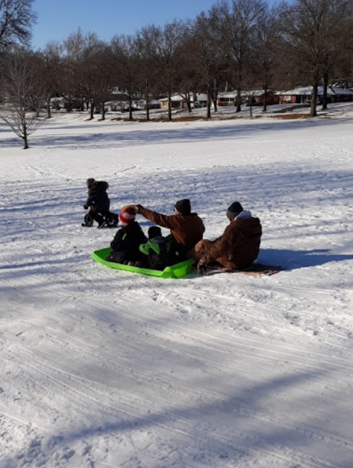Two people sledding down a snowy hill, while a child plays nearby in the snow. Trees and houses are visible in the background.