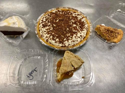 A variety of desserts on a table, including a chocolate pie, a cream pie, and slices of apple and crumb pie.