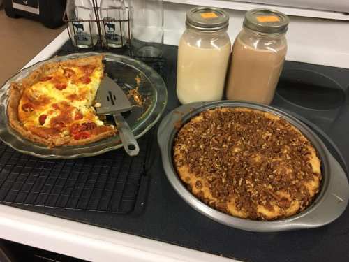 A savory pie with tomatoes and cheese next to a crumb-topped dessert pie and two jars of creamy beverages on a stove.