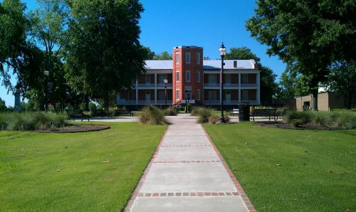 A pathway leads to a historic brick building surrounded by greenery and benches under a clear blue sky.