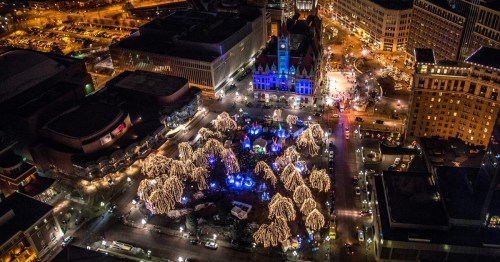 Aerial view of a city square illuminated with festive lights and decorated trees during the holiday season at night.