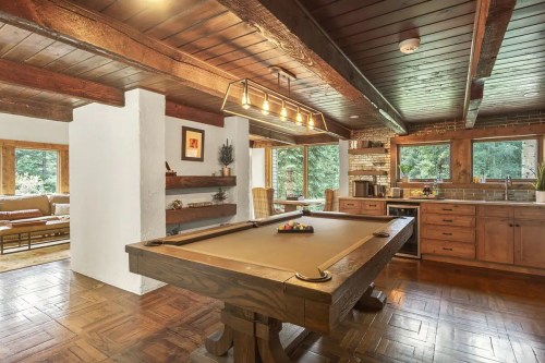Cozy interior featuring a pool table, wooden beams, and a kitchen with large windows overlooking greenery.