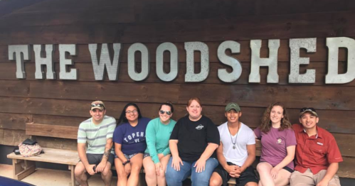 A group of seven people sitting in front of a wooden wall with the words "THE WOODSHED" displayed prominently.