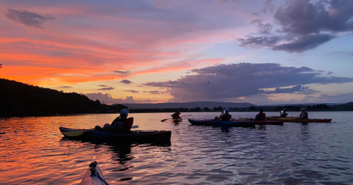 A group of kayakers on a calm lake at sunset, with vibrant orange and purple skies reflecting on the water.