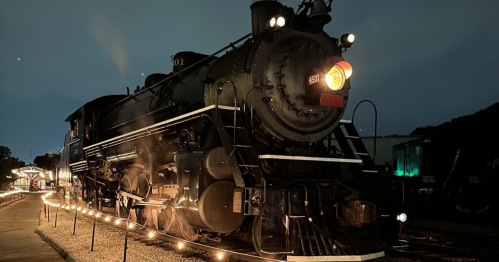 A vintage steam locomotive illuminated at night, with glowing headlights and steam rising from the engine.