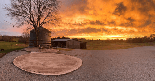 A vibrant sunset casts orange and yellow hues over a rural landscape with a silo, barn, and a tree.