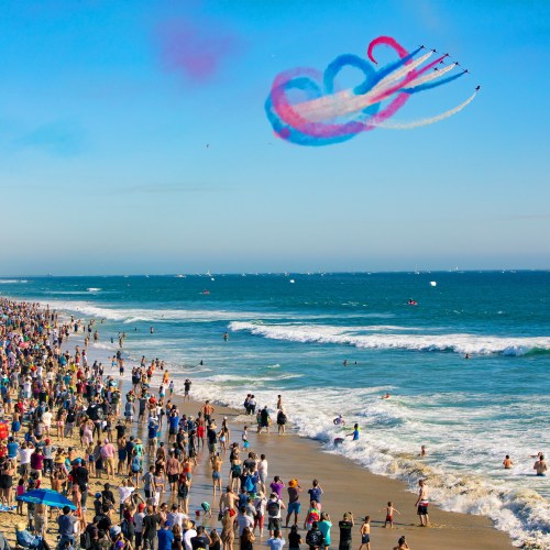 A crowded beach with people enjoying the sun, while colorful smoke trails from planes create patterns in the sky.