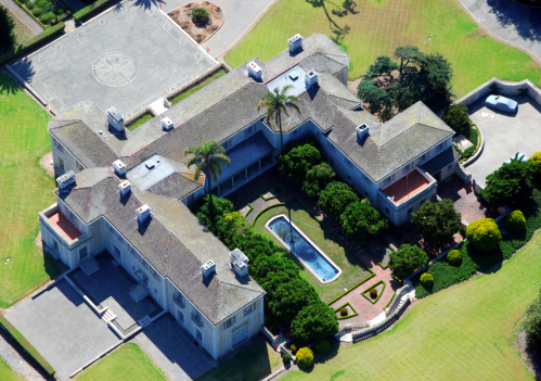 Aerial view of a large mansion with a pool and landscaped gardens, surrounded by green lawns and a paved area.