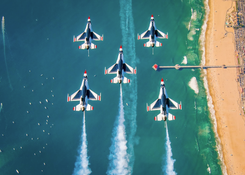 Five fighter jets flying in formation over a beach, leaving trails of smoke against a blue ocean backdrop.