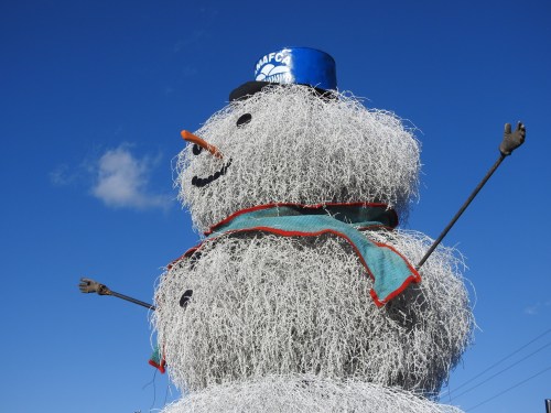 A large, whimsical snowman made of white material, wearing a blue hat and a scarf, against a clear blue sky.