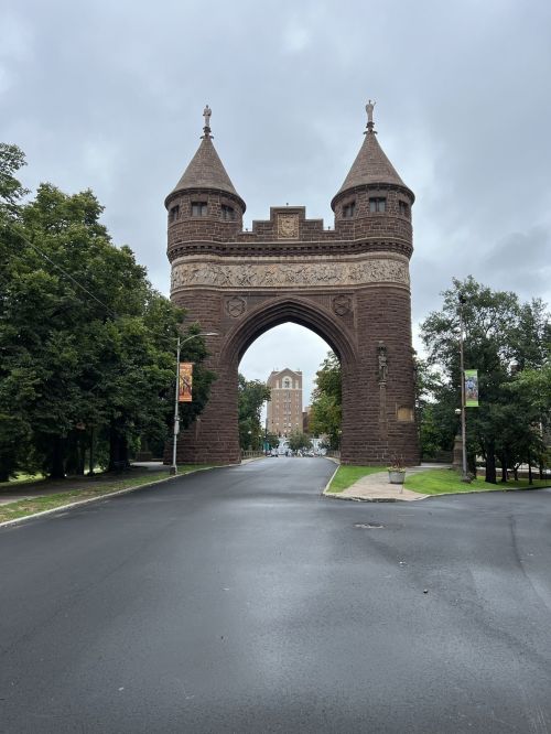 A stone archway with towers stands at the end of a tree-lined street on a cloudy day.