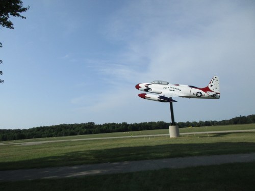 A vintage jet airplane sculpture on a pedestal, set against a clear blue sky and green landscape.