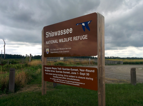 Sign for Shiawassee National Wildlife Refuge, detailing hours and trail information against a cloudy sky.