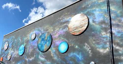 A colorful mural depicting planets and galaxies on a wall, with a blue sky and clouds in the background.