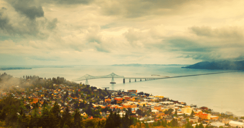Aerial view of a coastal town with a bridge, surrounded by water and lush greenery under a cloudy sky.