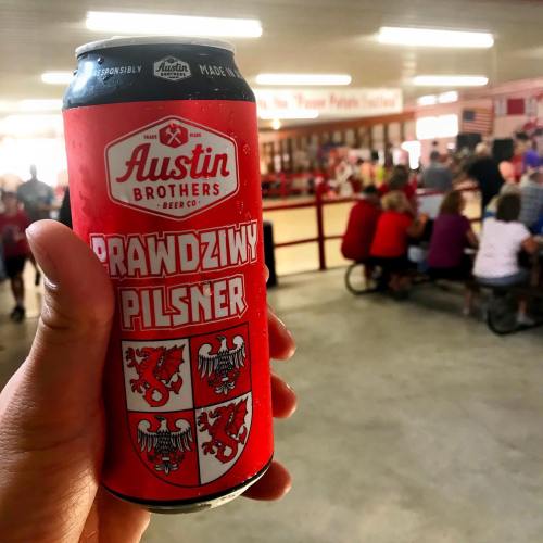 A hand holds a can of Austin Brothers Prawdziny Pilsner in a lively indoor setting with people in the background.