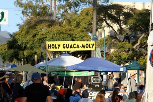 A lively outdoor event with a sign reading "Holy Guaca-Moly" and various tents, surrounded by a crowd of people.