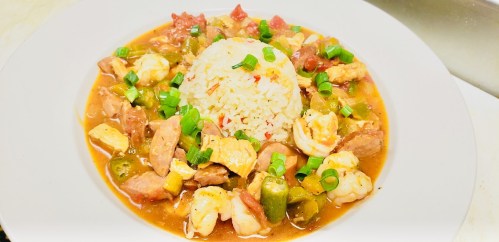 A bowl of gumbo with shrimp, sausage, and okra, served with a mound of rice and topped with green onions.
