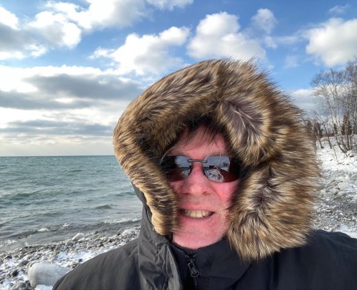 Person wearing a fur-lined hooded coat and sunglasses, standing by a snowy lakeshore with a cloudy sky.