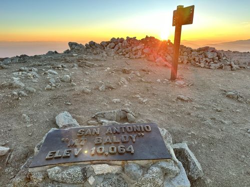 Sunrise over a rocky summit with a sign reading "San Antonio 'Mt Baldy' Elev. 10,064" in the foreground.