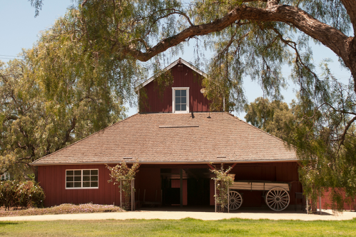 A red barn with a peaked roof, surrounded by trees and a wagon parked in front on a sunny day.