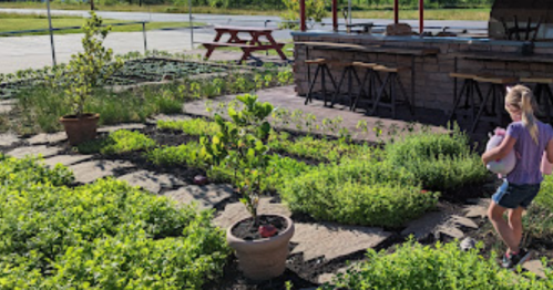 A child walks through a lush garden with potted plants and a picnic area in the background.