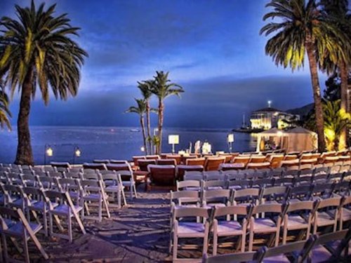 A serene beach setting at dusk with rows of white chairs and palm trees, overlooking calm waters and a distant building.