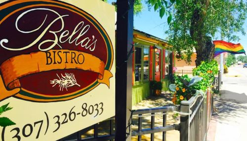 Sign for Bella's Bistro with colorful exterior and a rainbow flag nearby, surrounded by greenery and flowers.