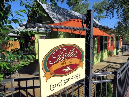 Sign for Bella's Bistro with a green building and orange umbrellas in the background, surrounded by trees.