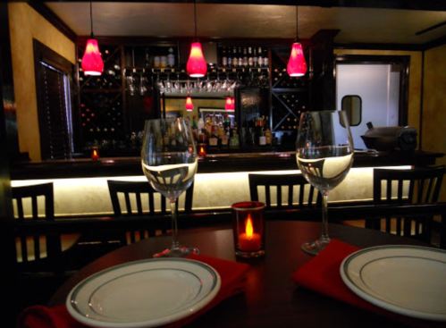 A cozy restaurant setting with two wine glasses, plates, and a softly lit bar in the background. Red pendant lights hang above.