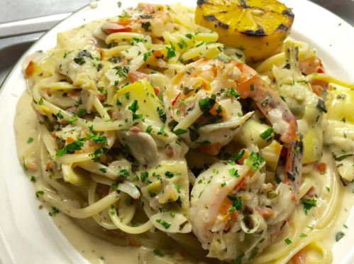 A plate of creamy pasta with shrimp, vegetables, and a grilled lemon slice, garnished with fresh herbs.