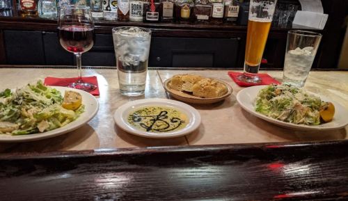 Two salads with lemon, drinks, and bread rolls on a bar counter with a variety of bottles in the background.